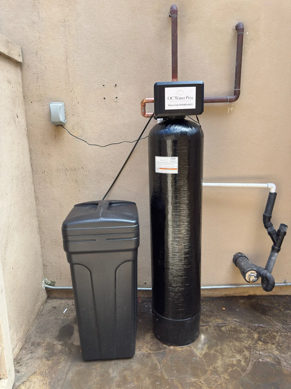 A single black softener tank and grey brine tank installed on a stucco exterior wall with copper piping