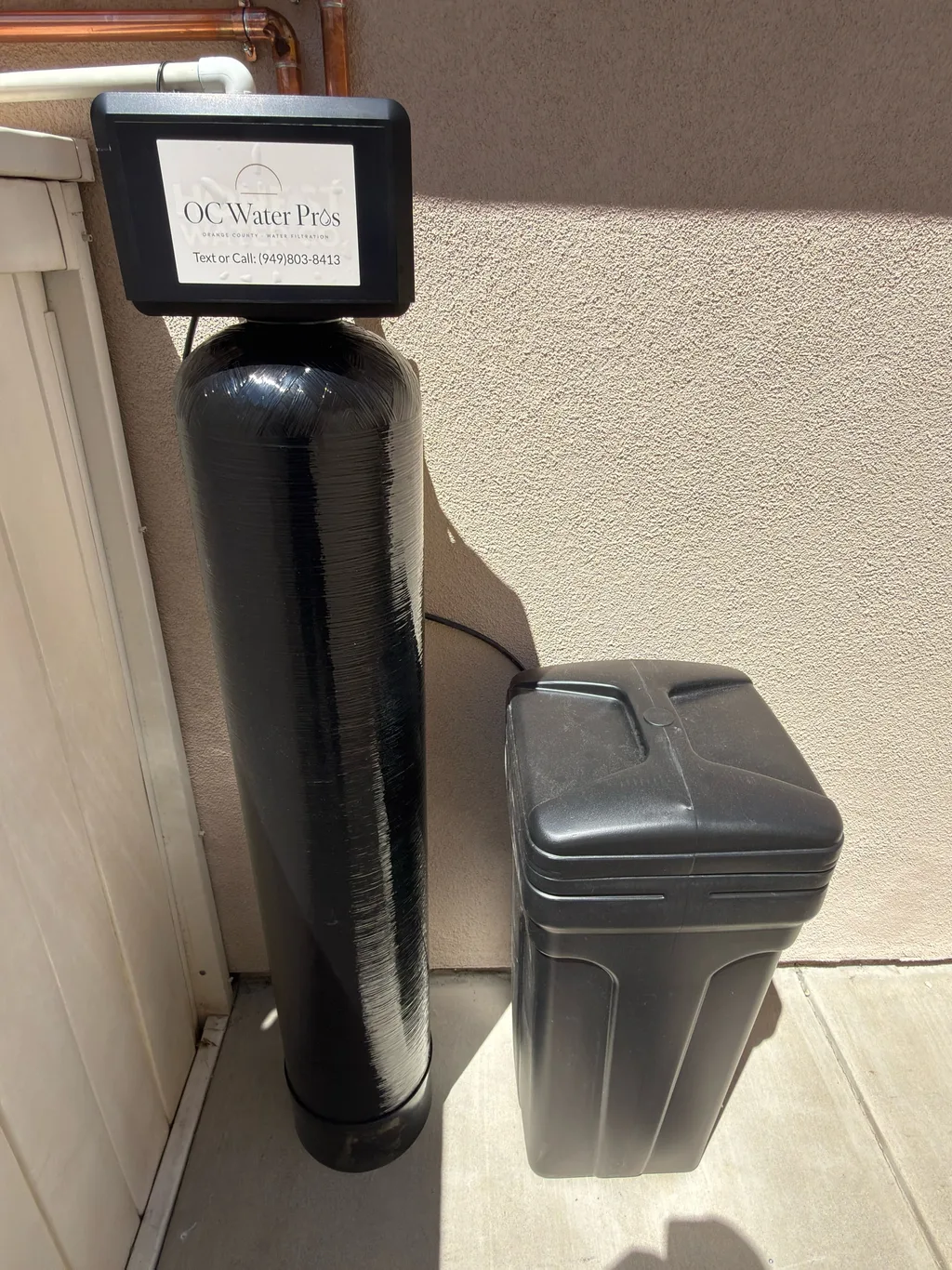 A black water softener tank and brine tank in a sunlit side walkway, OC Water Pros label on the valve head